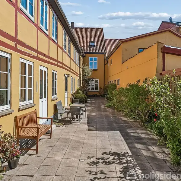 Gult bindingsværkshus med hvide vinduer, gårdhave med flisebelægning, træbænk og stole. Buske og planter langs siden. Solrigt vejr med blå himmel og spredte skyer.