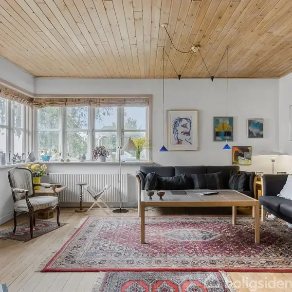 En stue med en sort sofa og sofabord, prydet af farverige billeder på væggene. Store vinduer lader lys komme ind, og tæpper dækker trægulvet. Møbler inkluderer stole og lamper.