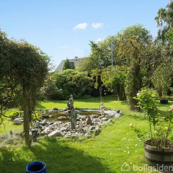 En have med en fuglefigur ved en klippeomkranset dam. Grønt græs og spredte tønder fyldt med blomster omgiver scenen, mens træer og buske danner en frodig baggrund under en klar blå himmel.