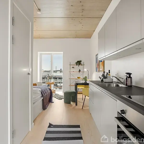 Moderne køkken med glatte, grå skabe og sort bordplade. Køkkenet strækker sig ind i en lys stue med en seng og skrivebord ved et vindue. Striped tæppe på gulvet.