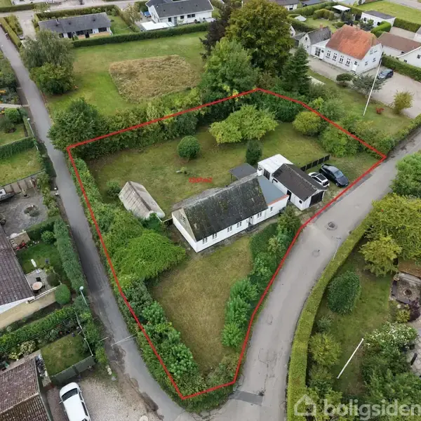 Hus set oppefra med have og garage inden for rød markering. Huset ligger ved vejkryds omgivet af andre boliger og grønne områder.