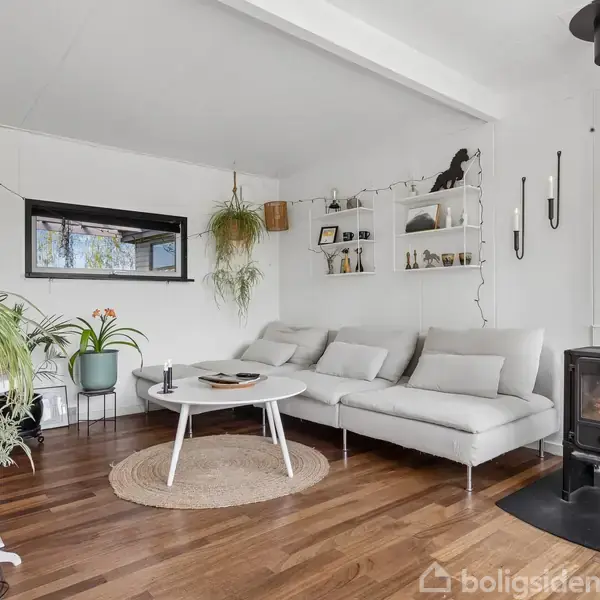 Stue med en lys sofa, der står ved siden af en moderne brændeovn, som er i brug. Rummet har trægulv, mange planter og hylder på væggen.