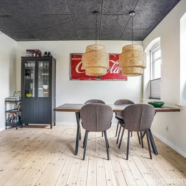 Et spisebord med fire grå stole står i et lyst rum. To loftslamper hænger over bordet. På væggen er et rødt skilt med teksten "Original Coca-Cola."