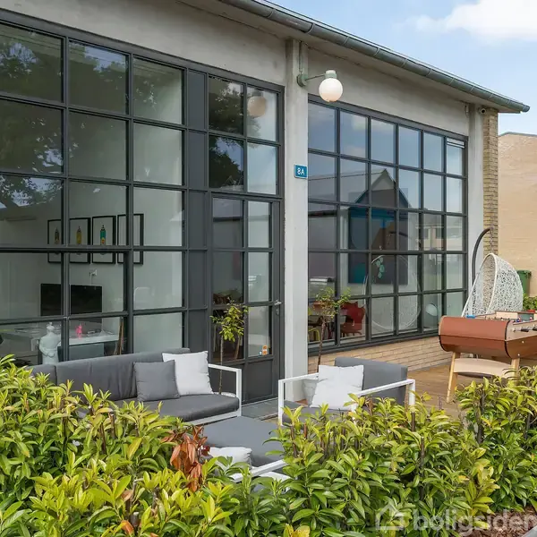 Moderne terrasse med grå sofasæt og grill foran bygning med store glasvinduer. Omkring er der frodige buske og grønne planter.