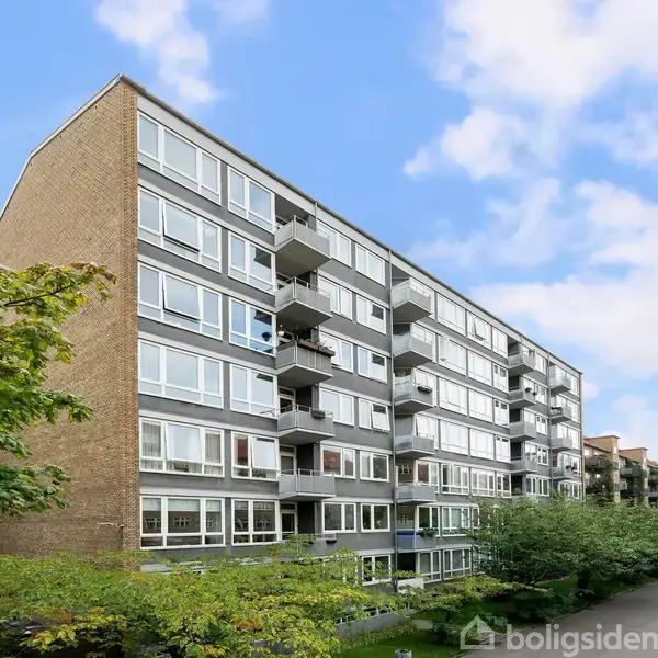 Etagebygning med altaner strækker sig mod himlen omgivet af grønne træer i et byområde. Bygningen har mange vinduer og er placeret foran en flisebelagt sti under en blå himmel.