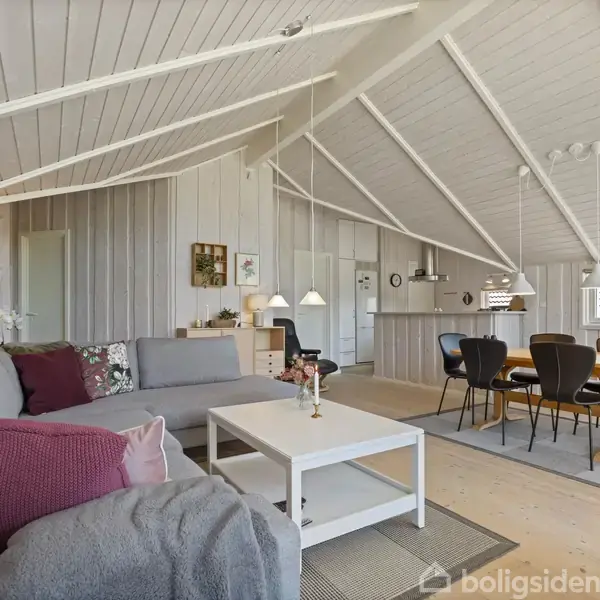 Stue med grå sofa og hvide sofabord, udsmykket med røde puder; spisebord med sorte stole i baggrunden. Loftets træbjælker skaber en hyggelig atmosfære i rummet.