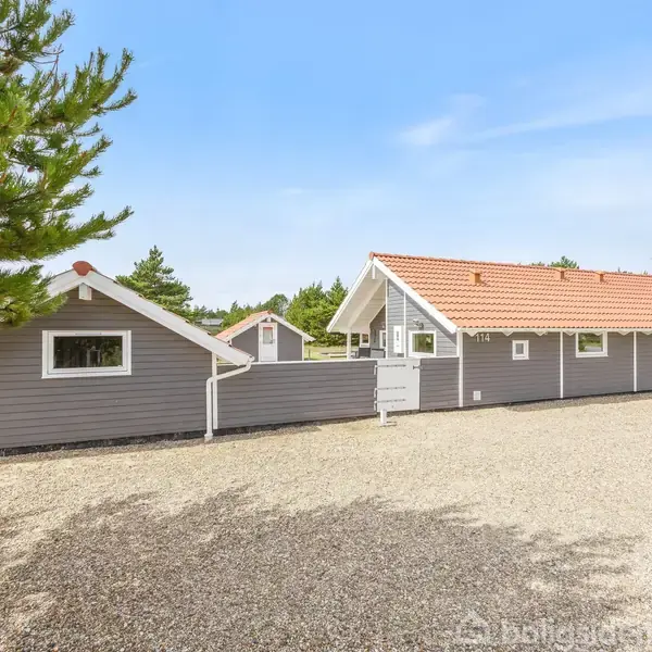 Gråt hus med rødt tag står på en grusplads omgivet af træer. Ved siden af er en mindre bygning. Klar, blå himmel i baggrunden.
