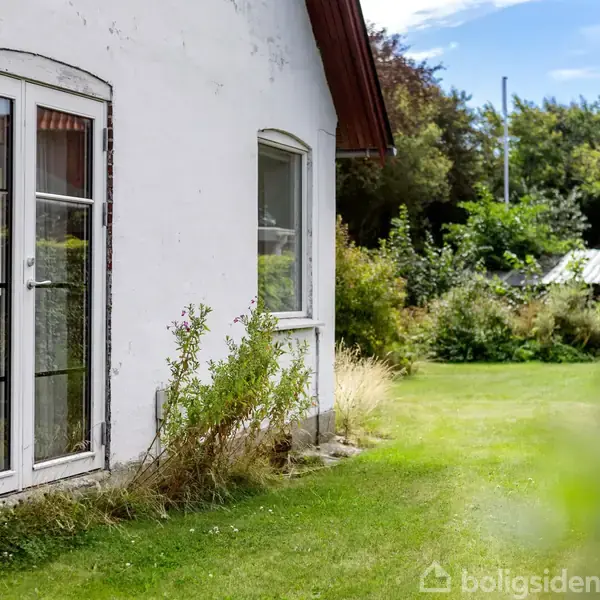 Et hvidt hus med glasdør og vindue står i en have med grønt græs og buske. Solen skinner, og der er træer i baggrunden.