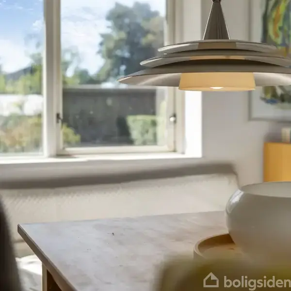 En moderne pendellampe hænger over et træbord i et lyst rum med en stor vindue med udsigt til natur. En skål og bøger er på bordet og hylden.