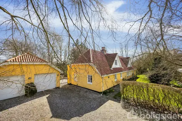 Gul villa med rødt tag står stille omkranset af træer og grønne buske. I forgrunden grusbelagt indkørsel ved siden af en garage. Solrigt og klart vejr.