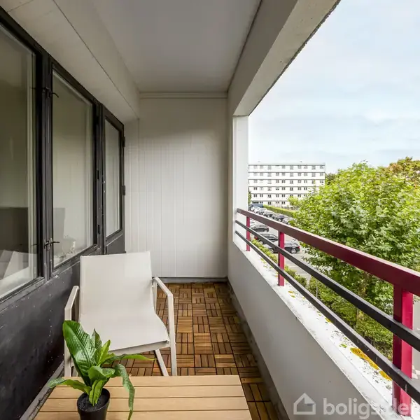 En balkon med træfliser og en hvid stol ved et bord med en plante. Der er udsigt over grønne træer og en lejlighedsbygning i baggrunden.