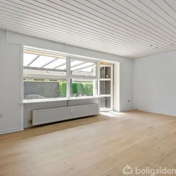 En tom stue med lyst trægulv har et stort vindue og glasdør til højre, der åbner mod en udestue. Rummet er minimalistisk med hvide vægge og loft.