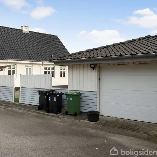 En garage med hvid og grå facade står ved siden af en villa med sort tag. Affaldsspande er placeret ved siden af garagen. Himlen er blå og skyet.