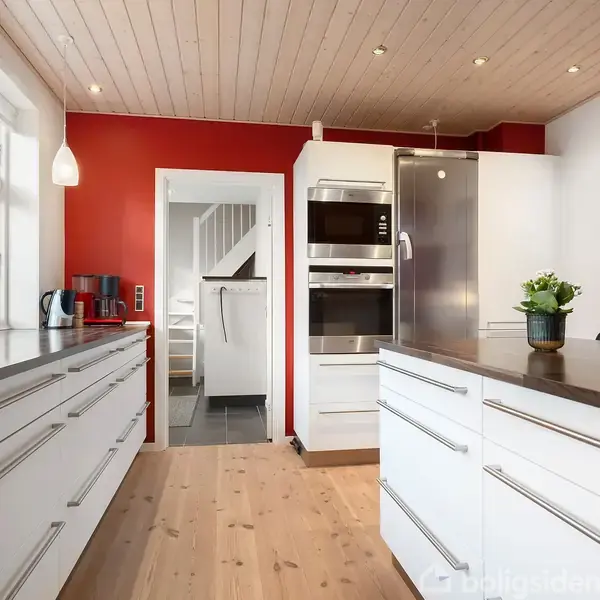 Et moderne køkken med hvide skabe og sorte bordplader. Køleskab og ovne er bygget ind i væggen. Rummet har trægulv, røde og hvide vægge, og store vinduer med naturligt lys.