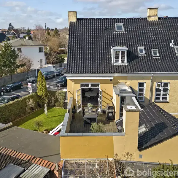 En gul murstensbygning med sort tegltag har en terrasse med havemøbler. Omkring er der en velplejet græsplæne og parkerede biler på vejen i et villakvarter.