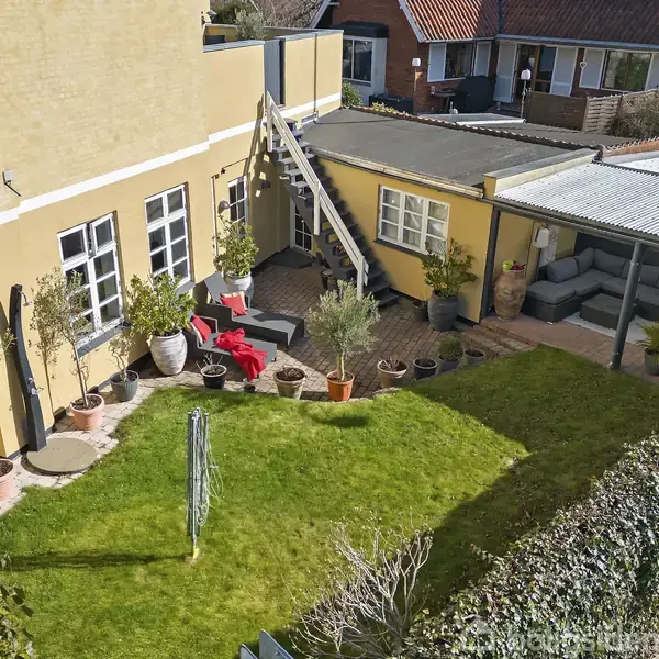 Gul bygning med trapper op til taget, omgivet af en grøn græsplæne og flere potteplanter. Ved siden af bygningen er der en terrasse med sofa og planter.