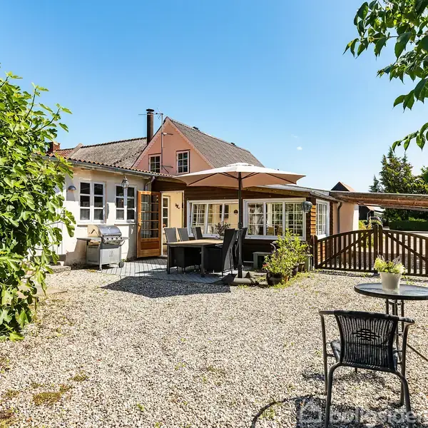 En grusbelagt gårdhave med et bord og stole under en parasol. Omkring er buske, et hegn, og et hus med store vinduer. Solrig himmel i baggrunden.