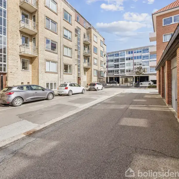 Bygninger står langs en stille gade, hvor biler er parkeret. Solen skinner, og en moderne lejlighedskompleks ses i baggrunden med glasfacader. En cykel kan også skimtes til venstre.