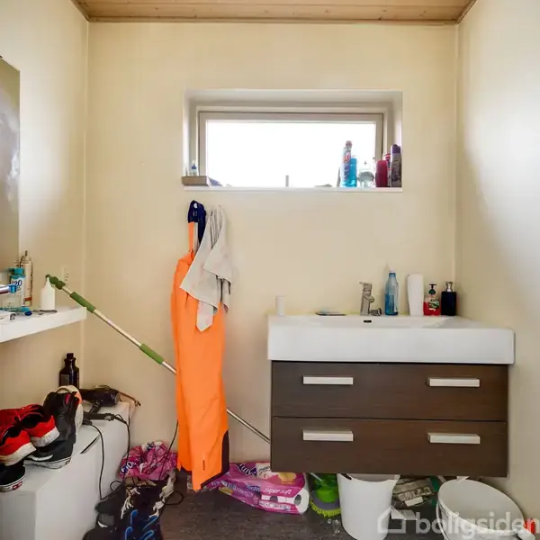 Et badeværelse med en håndvask og skab i midten omgivet af diverse toiletartikler. Til venstre står rengøringsartikler og sko, mens der hænger orange arbejdstøj på en krog. En lille vindue over vasken.