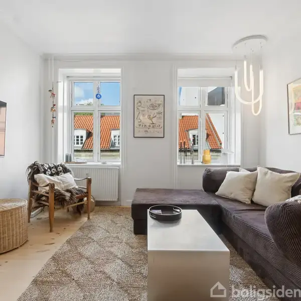 En stue med en mørk L-formet sofa står ved siden af et moderne sofabord og en flettet stol. Store vinduer giver udsigt til røde hustage, omgivet af malerier og et tæppe på gulvet. En fladskærm hænger på væggen.
