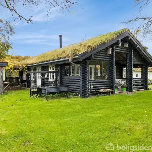 Træhus med græstag står stille på en grøn græsplæne omgivet af træer og buske, ved siden af en trægarage. Skyfri blå himmel ses i baggrunden.
