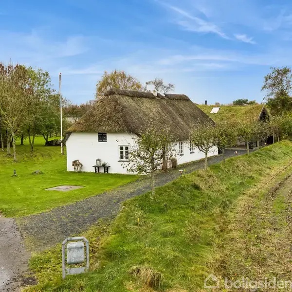Hvidt stråtækt hus, omgivet af grønne træer, står stille langs en landevej ved siden af en pløjet mark. Blåt himmel og vejskilt kompletterer det landlige landskab.