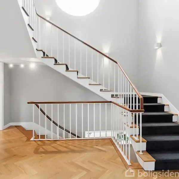 Trappeopgang med hvidmalede gelændere og sorte trappetrin. Trappen er i en lys entre med parketgulv og væglamper.