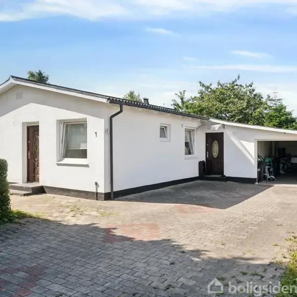 En hvid bungalow med fladt tag står i solskin. Den har en stor indkørsel foran og en garage til højre. Flere buske og træer omgiver huset.