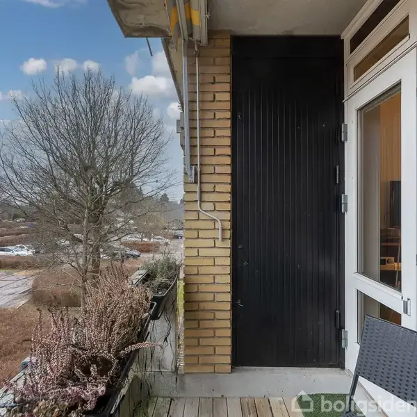 Dør åben i en murstensvæg ud til en altan med træplankegulv. Omkring ses nøgne træer, parkerede biler og en overskyet himmel. Stole nær døren.