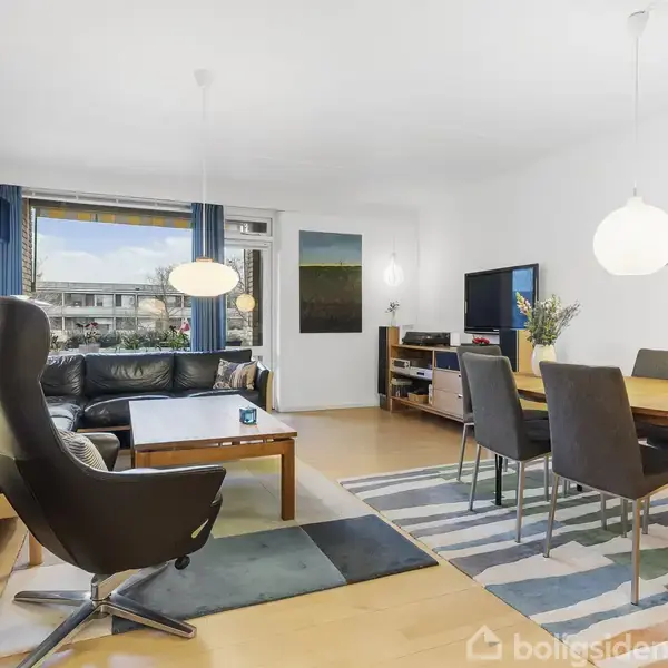 Stue med sorte lædermøbler og træbord i midten, flankeret af spisestuemøbler og fladskærms-TV. Vindue med blå gardiner til venstre, malerier på væggene. Skaber lys og moderne stemning.