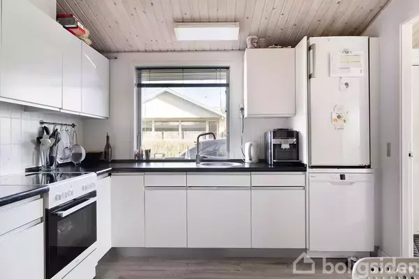 Et moderne, lyst køkken med hvide skabe og sorte bordplader. Køleskab, ovn og opvaskemaskine er integreret. Et vindue med udsigt til en terrasse dominerer væggen over vasken.