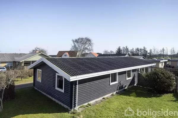 Et sort træhus med hvide vinduesrammer står stille i en have omgivet af andre huse. Græsplænen er velplejet, og træer er synlige i baggrunden under en blå himmel.