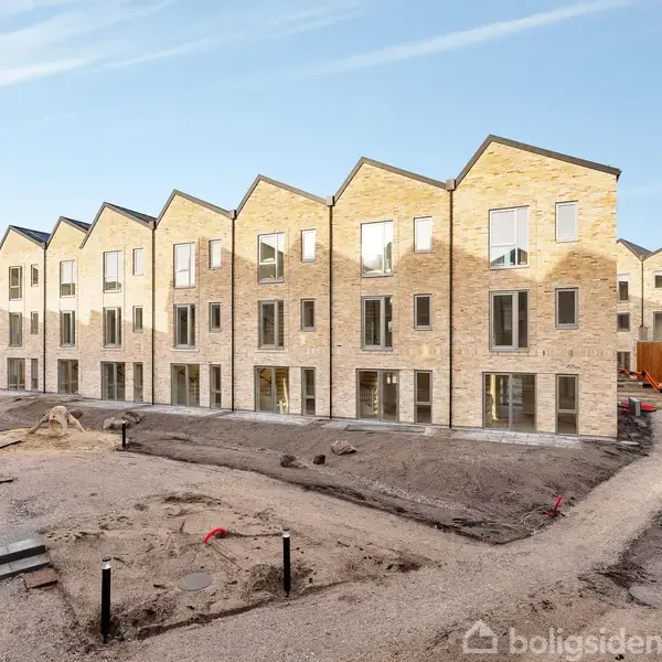 Rækkehuse i lyse mursten står i række. Ingen bygger eller arbejder, omgivelserne præget af byggeplads med jord og gangstier. Skyfri himmel giver et klart lys.