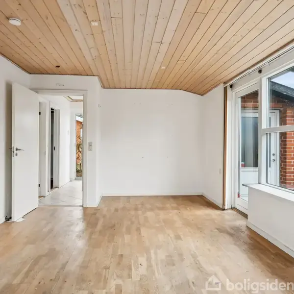 Et lyst rum med trægulv og træloft har åbne døre til en gang. Store vinduer til højre viser udsigt til en murstensbygning og tillader naturligt lys at trænge ind.