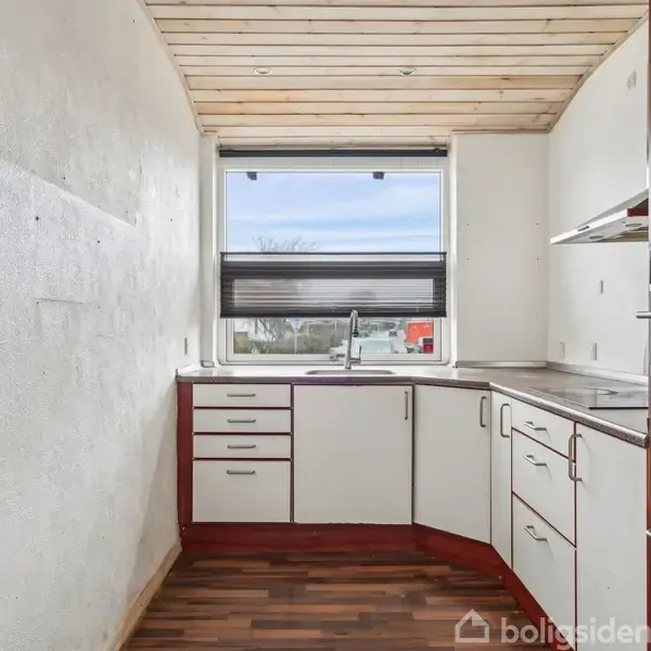 Et lille køkken med hvide skabe og en brun bordplade. Der er en vask under et vindue med udsigt til himmel. Trægulv og lofter, indbygget ovn til højre.