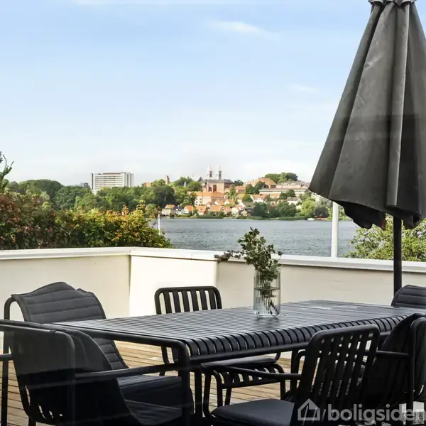 En terrasse med et bord og fem stole står under en stor parasol. I baggrunden ses en udsigt over vandet til en by med grønne områder og bygninger.