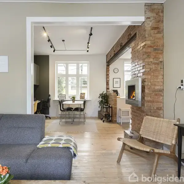 En stue med grå sofa og træstol, hvor en moderne pejs er indbygget i en murstensvæg. Spisestuen i baggrunden har et bord ved et vindue, og trægulve gennem hele rummet.