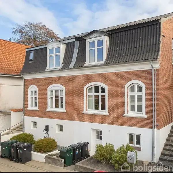 Et rødt murstenshus med sorte tagsten står i et boligområde, omgivet af andre huse. De har buede hvide vinduer og flere skraldespande foran.