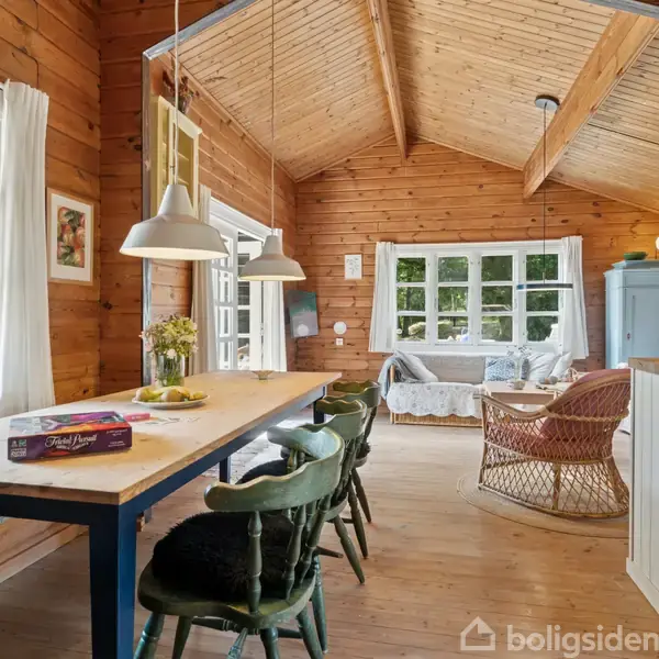 Træspisebord med stole står centralt, omgivet af trævægge og store vinduer. Lamper hænger over bordet. I baggrunden ses en sofa og skrivebord nær et blåt skab. Ingen tekst.