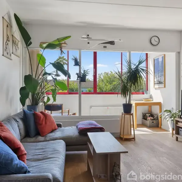 Sofaen står i en stue med planter og farverige puder, vendt mod store vinduer med udsigt til træer. Vægge har plakater, og et ur hænger over vinduerne. Bord og TV.
Mamma Andersson-plakat.