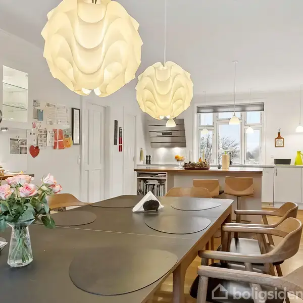 Et moderne køkken med et stort spisebord og træstole. Bordet har en vase med blomster. Stemningsfulde lamper hænger ned, og der ses hvide skabe og apparater i baggrunden.