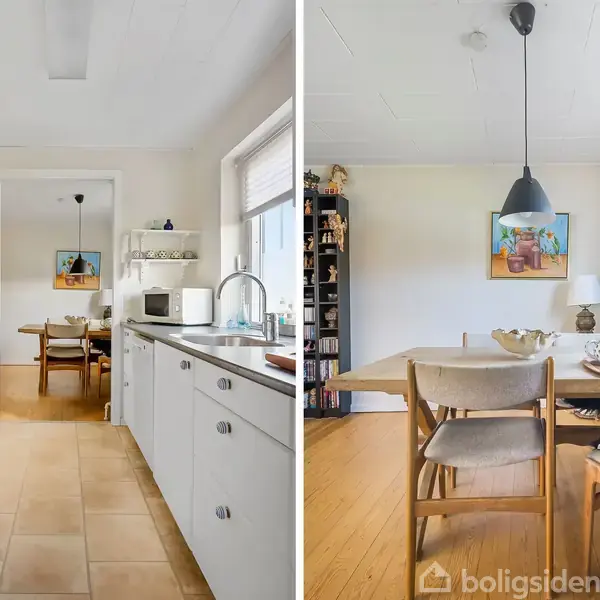 Køkken med hvide skabe og grå bordplade, set med udsigt til spisestue med træmøbler, hængende lampe og billeder på væggen. Lyst rum med vindueslys og planter.
