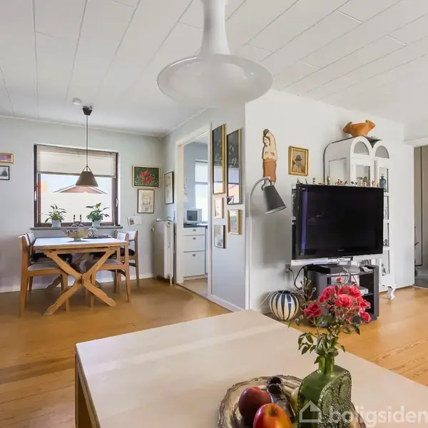 Et moderne opholdsrum indeholder en sofabord med frugt og blomster i forgrunden, en reol med fladskærms-tv til højre, spisebord i baggrunden. Rummet har trægulv og er lyst.