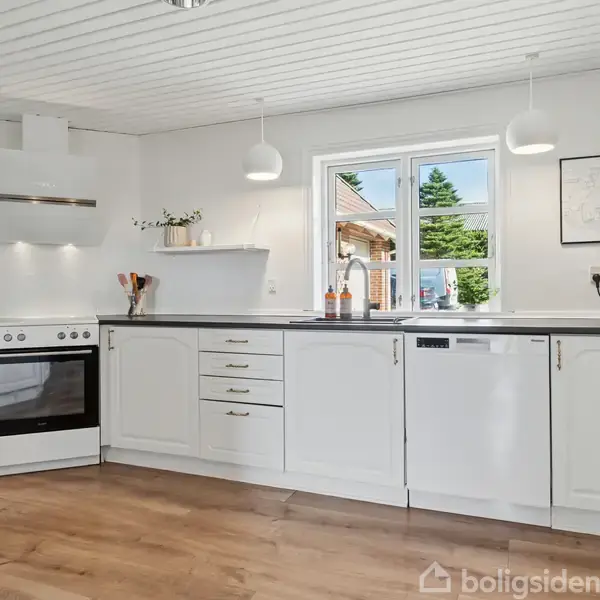 Et moderne køkken med hvidt interiør og trægulv. Køkkenet indeholder en komfur med emhætte, opvaskemaskine, knivblok, og planter på hylde. Vindue viser udsigt til en gårdhave.