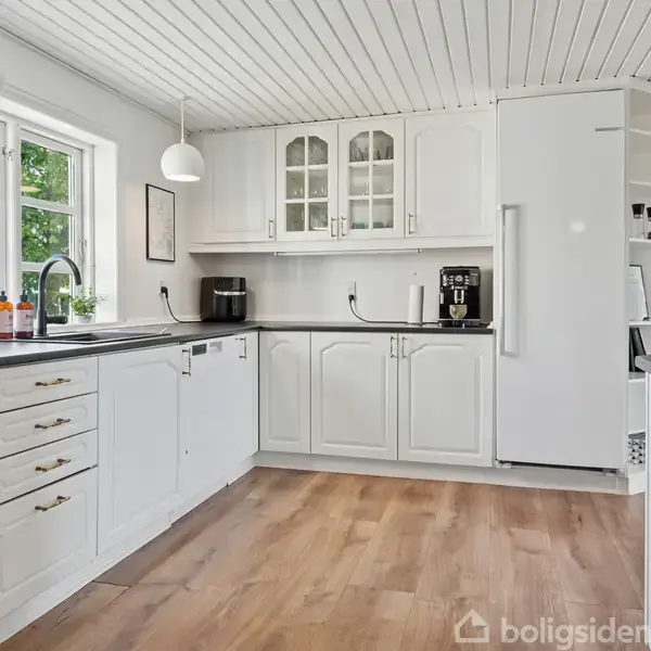 Hvidt køkken med lysebrune trægulve. Skabe og skuffer er lukket, mens kaffemaskinen og blenderen står på sort bordplade. Vindue med udsigt til grøn natur.