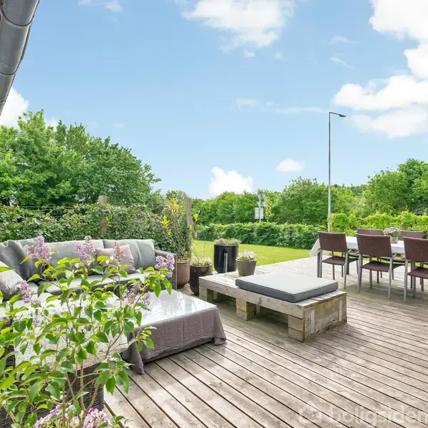 Terrassen har to siddeområder, et med sofa og bord og et med spisebord og stole, omgivet af grønne planter under en klar blå himmel.