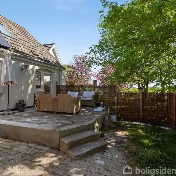 Terrasse med møbelsæt og solskærm. Møblerne står oven på flisebelægning foran et hus med solpaneler på taget. Omkring er der grønne træer og en flethegn.