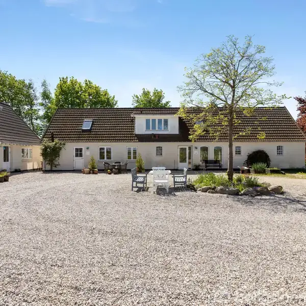 Hvidt, udstrakt hus med mørkt tag står i en grusbelagt gårdsplads. En lille havemøbelgruppe og et træ befinder sig i midten. Omkranset af grønne træer.