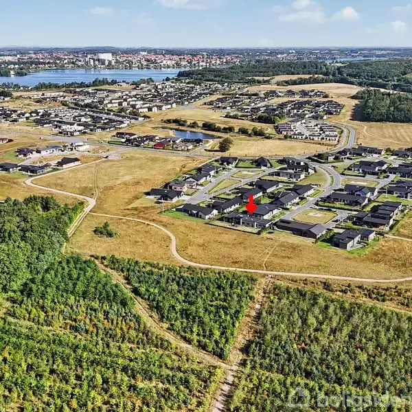 Et boligområde ligger i et åbent landskab omgivet af skove og græsarealer. Huse er arrangeret langs snoede veje. I baggrunden ses en sø og en by.