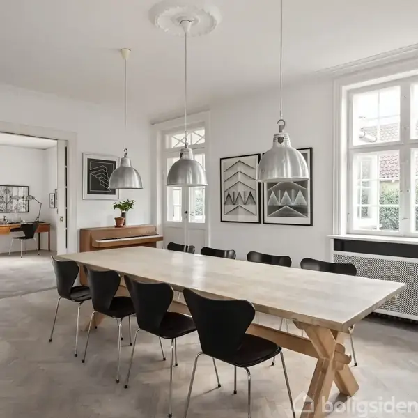 Et langt træbord med sorte stole står i en lys spisestue. Tre metallamper hænger over bordet. Der er kunst på væggene, og rummet har store vinduer med udsigt til en gårdhave.